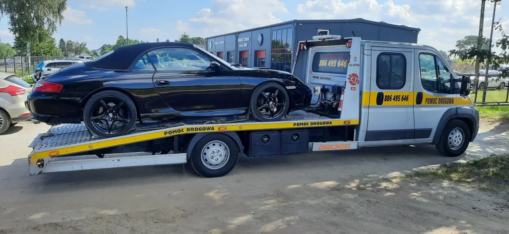 Ujęcie z boku (profil) na lawetę pomocy drogowej ze Słupska z czarnym autem na platformie. Pojazd stoi na piaszczystej drodze przed budynkiem warsztatu samochodowego.