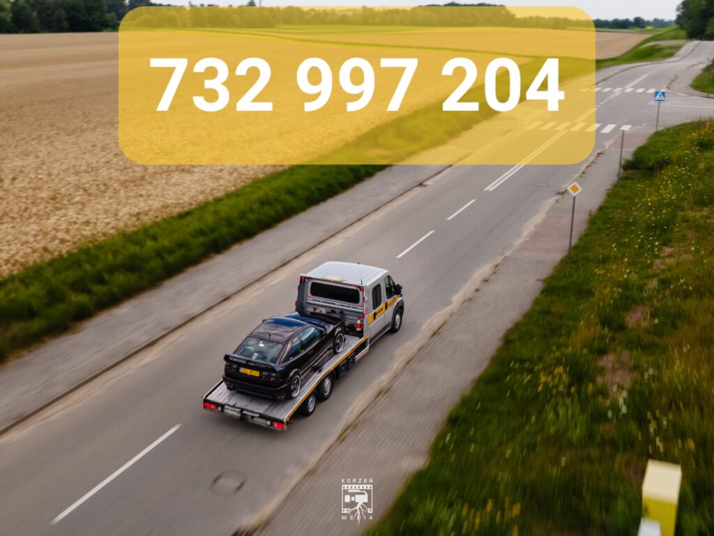 Widok z lotu ptaka na lawetę pomocy drogowej ze Słupska, jadącą tyłem do kamery w stronę skrzyżowania. Po lewej stronie widać pola, a na górnym żółtym pasku widnieje biały numer telefonu