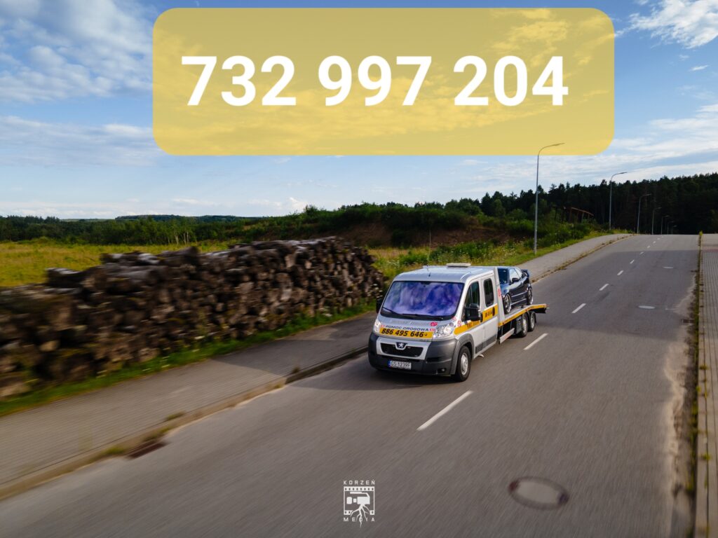 Widok z przodu na lawetę pomocy drogowej ze Słupska, jadącą drogą krajową z autem na platformie. W tle widać drzewa i ułożone na poboczu stosy drewna. Na górze zdjęcia, na żółtym tle, widnieje numer telefonu