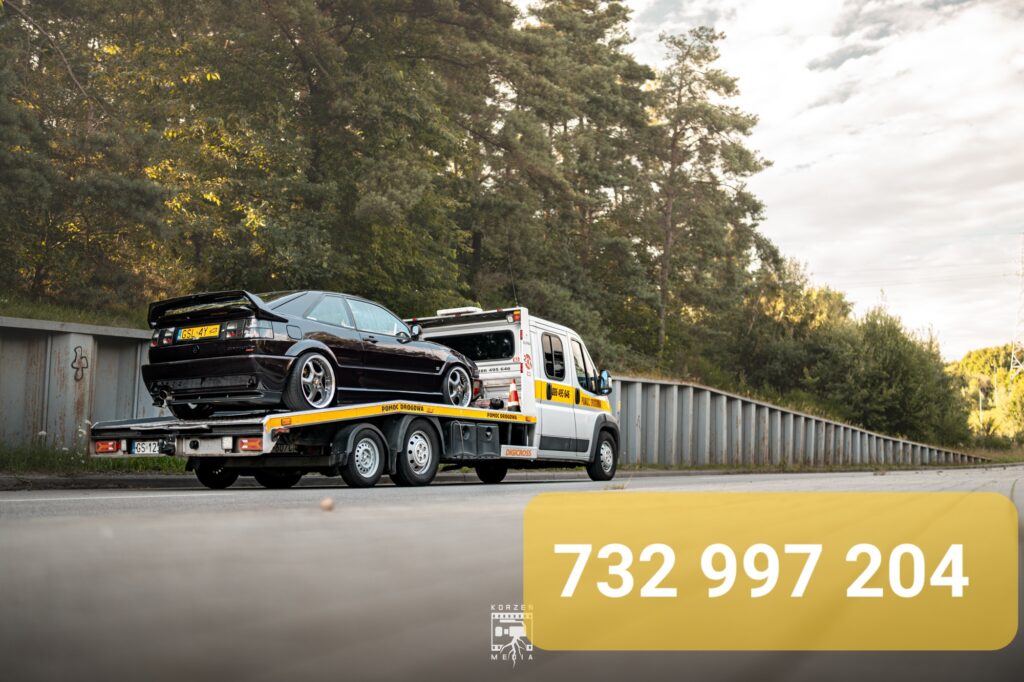 Laweta pomocy drogowej ze Słupska widziana pod kątem, z widocznym tyłem transportowanego samochodu na platformie. W tle widać korony drzew, a na dolnym żółtym pasku widnieje numer telefonu