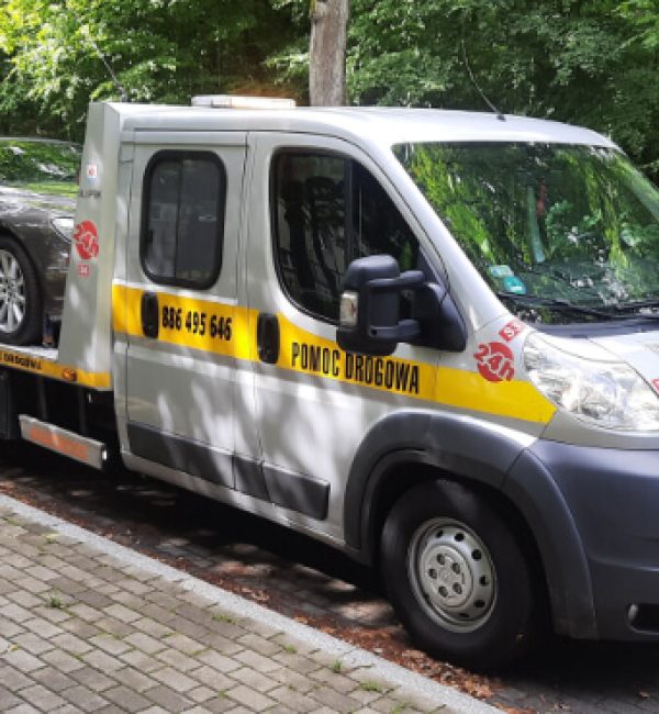 Widok z przodu pod lekkim kątem na lawetę pomocy drogowej ze Słupska z czarnym samochodem na platformie. Pojazd stoi w zatoczce z kostki brukowej na tle drzew.