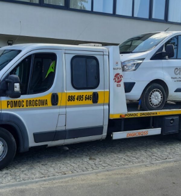 Laweta pomocy drogowej ze Słupska podczas transportu białego auta dostawczego z osiedlowego parkingu. Pojazd stoi na kostce brukowej, a w tle widać blok mieszkalny; ujęcie pod lekkim kątem z boku.